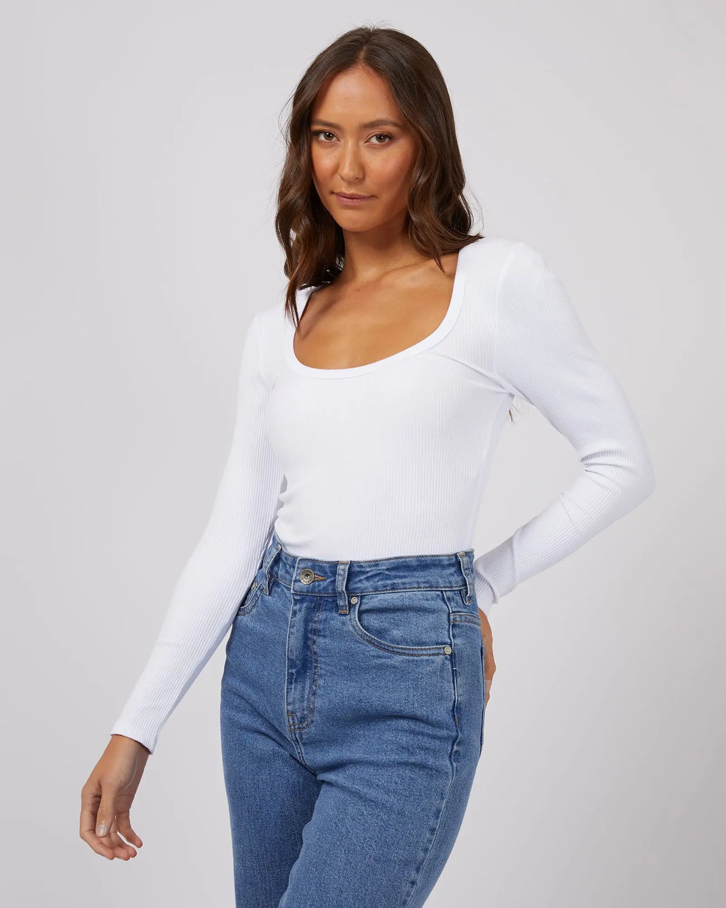 Woman wearing a white long-sleeve top and blue jeans on a plain background