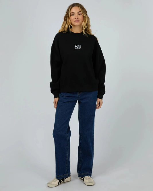 Person wearing a black sweatshirt with a logo and blue jeans on a gray background