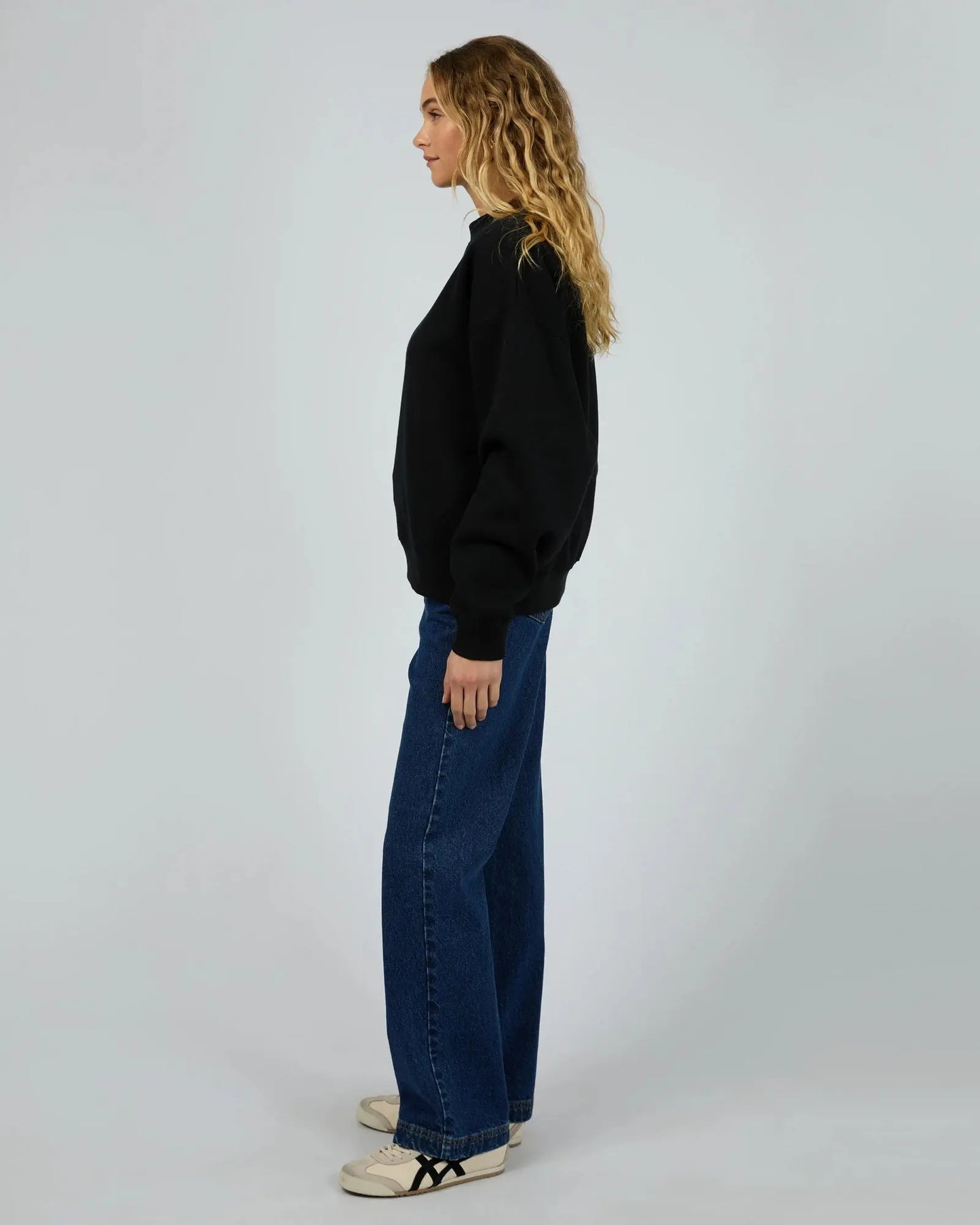Person wearing a black sweater and blue jeans standing against a plain background