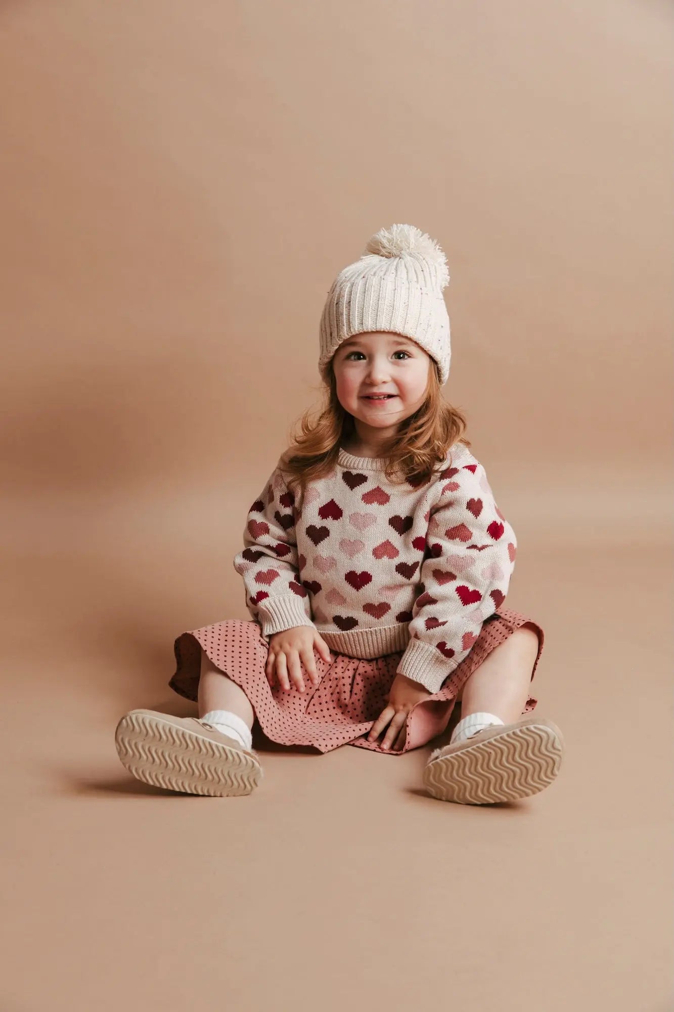 Child wearing a heart-patterned sweater and white knit hat on a beige background