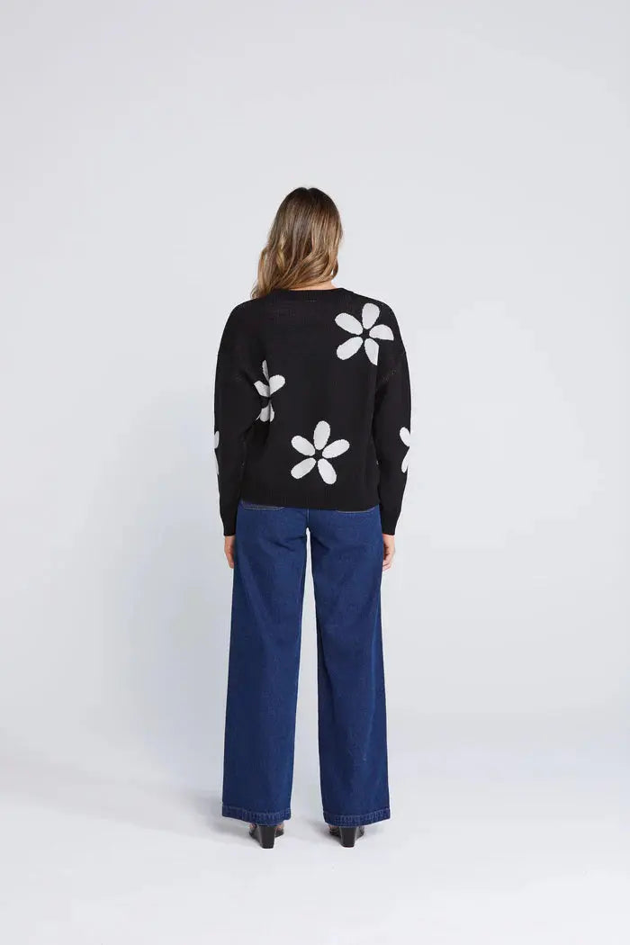 Person wearing a black sweater with white floral patterns and blue pants on a white background