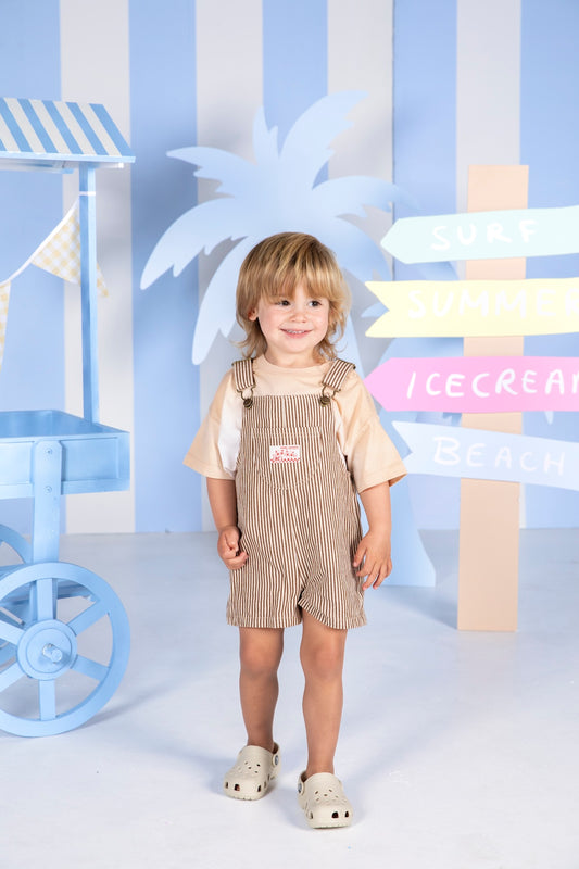 A young child wearing beige and white vertically striped overall shorts over a light beige t-shirt, standing in a beach-themed studio setting with blue and white striped walls, a palm tree cutout, and colorful signpost decorations.