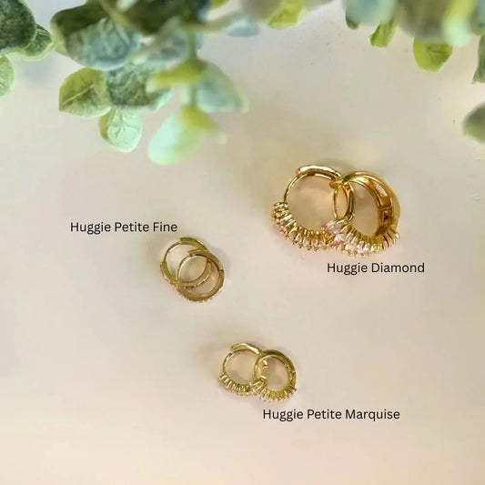 Three gold hoop earrings labeled 'Huggie Petite Fine', 'Huggie Diamond', and 'Huggie Petite Marquise' on a light background with green leaves.