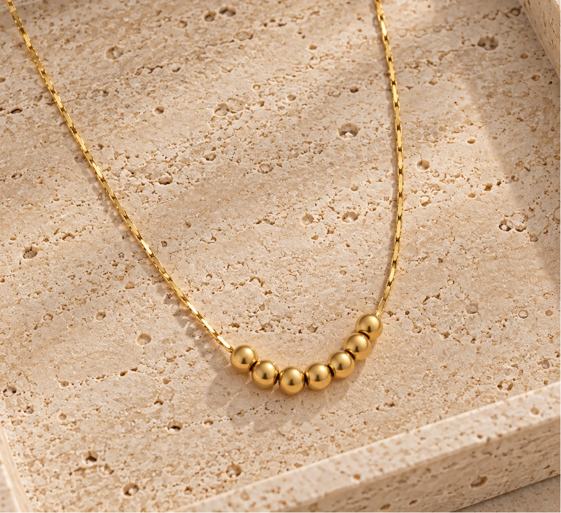 A delicate gold chain necklace with a curved row of small polished gold beads, displayed on a textured beige stone tray.