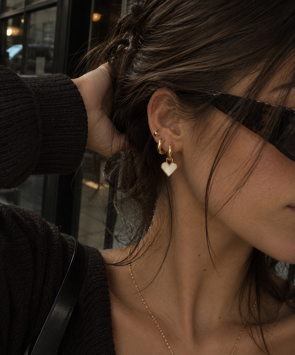 A close-up lifestyle shot of a woman wearing a pair of gold double-hoop earrings with a small white heart dangle, styled with sunglasses and a gold chain necklace.