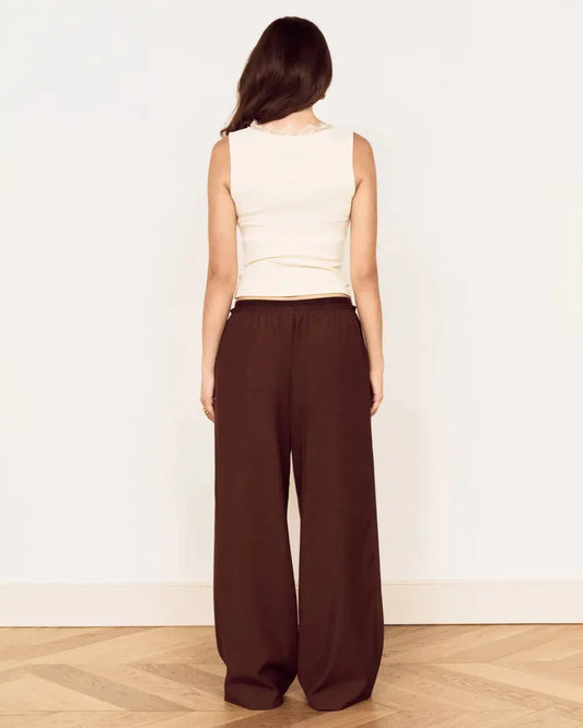 A woman shown from the back wearing a cream sleeveless top and dark brown wide-leg pants, standing on a light wood herringbone floor against a plain white wall.