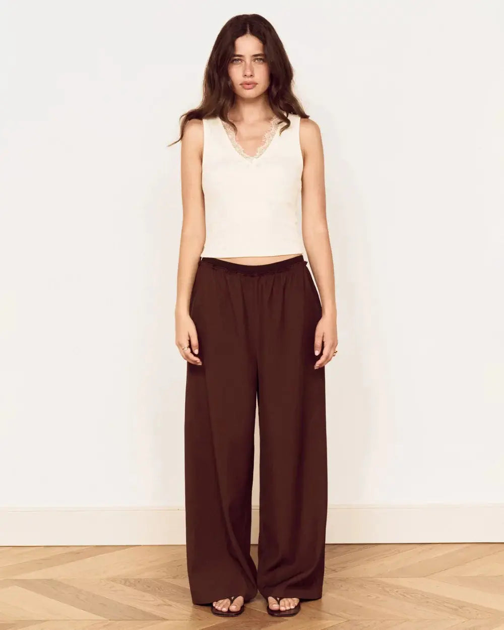 A woman stands against a plain white wall wearing a sleeveless cream top and dark brown wide-leg pants, with sandals on a light wood floor.