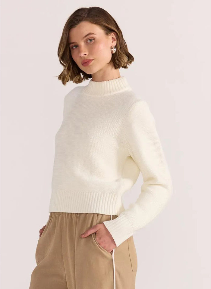 Woman wearing a white sweater and beige pants against a light background