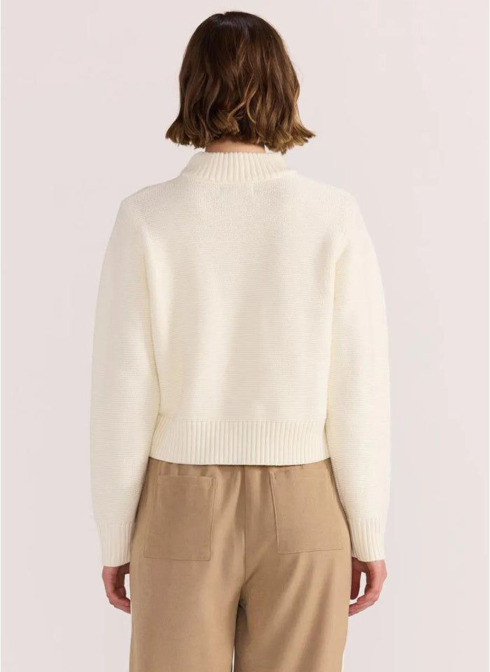 Person wearing a cream sweater and beige pants on a light pink background