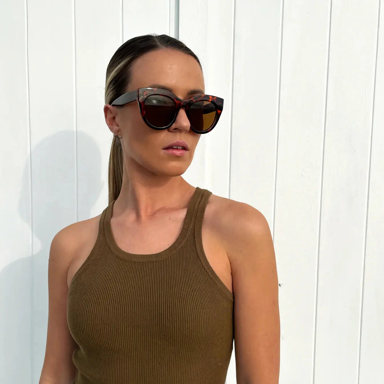 Woman wearing sunglasses and a brown tank top against a white wooden background