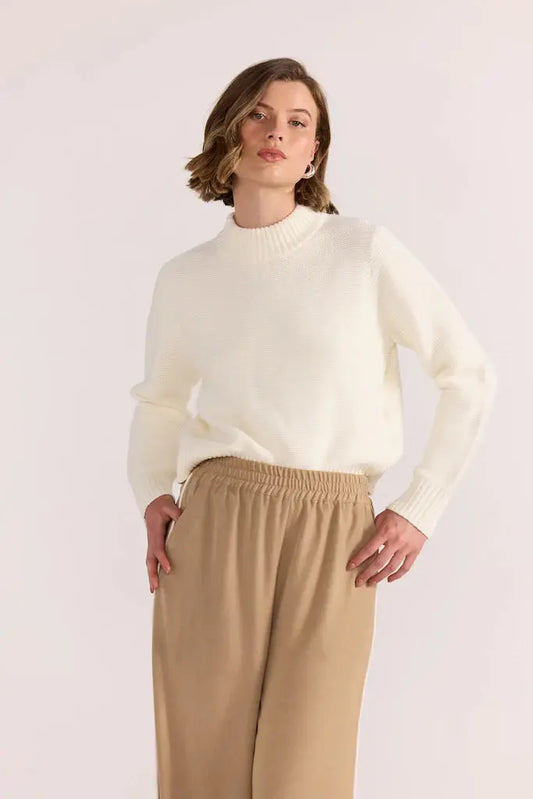 Woman wearing a cream sweater and beige pants against a white background