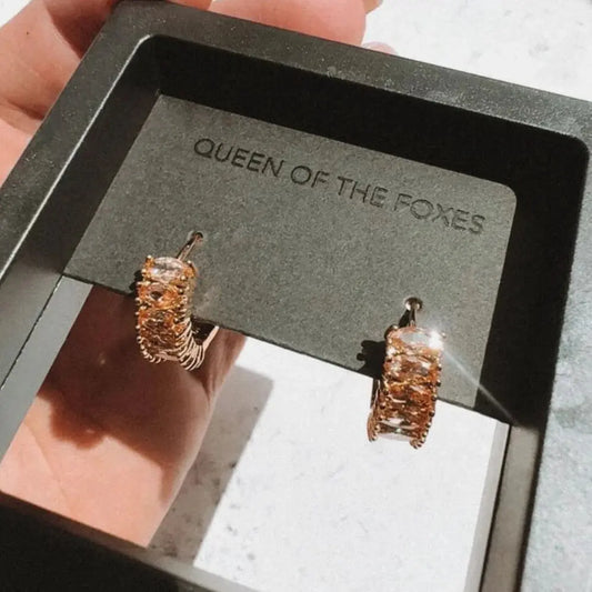 Gold earrings on a 'Queen of the Foxes' branded card held by a hand. The Perfect Huggies Diamond - The Rose Edit