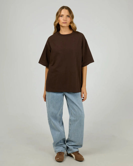 Person wearing a brown oversized t-shirt and blue jeans on a plain background. Core Oversized Tee Chocolate - The Rose Edit
