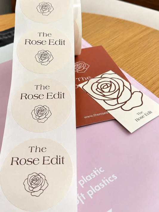 A roll of The Rose Edit custom matte stickers resting on tissue paper packaging inserts and a postage bag