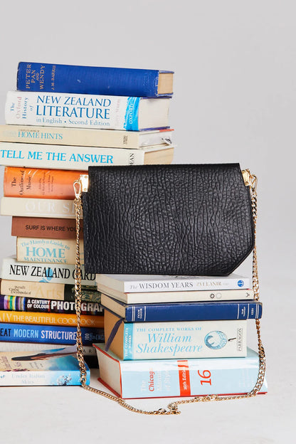 Black textured handbag with gold chain over a stack of books on a light gray background