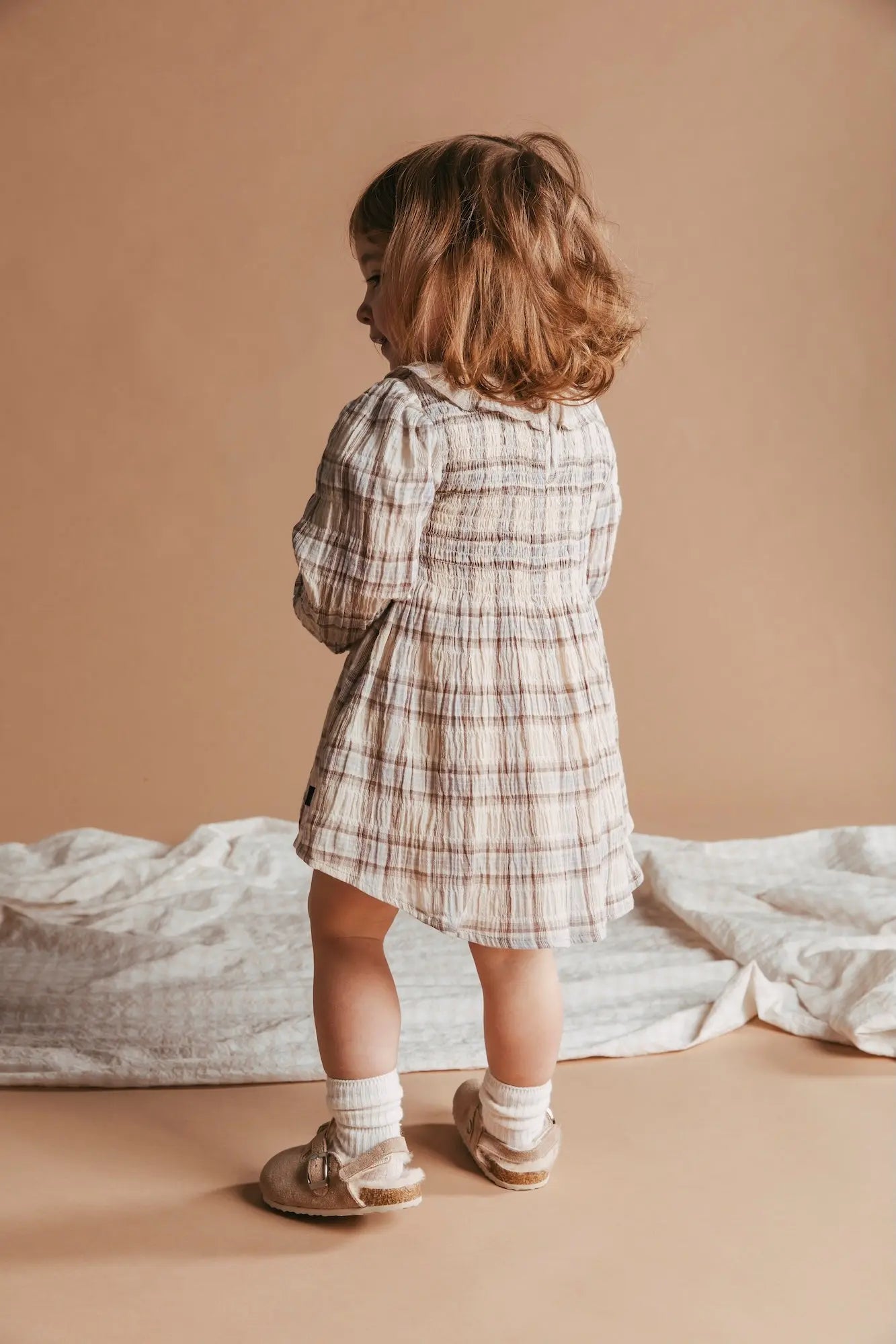 Child wearing a plaid dress standing on a beige floor with a neutral background