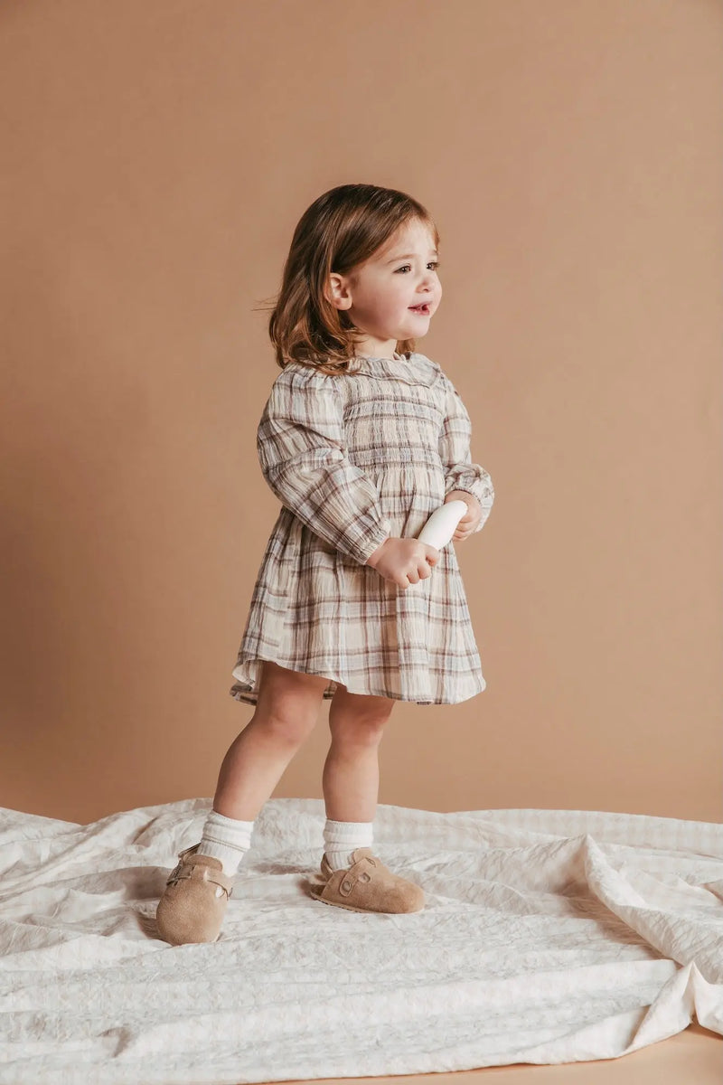 Young child wearing a plaid dress standing on a beige blanket against a beige background