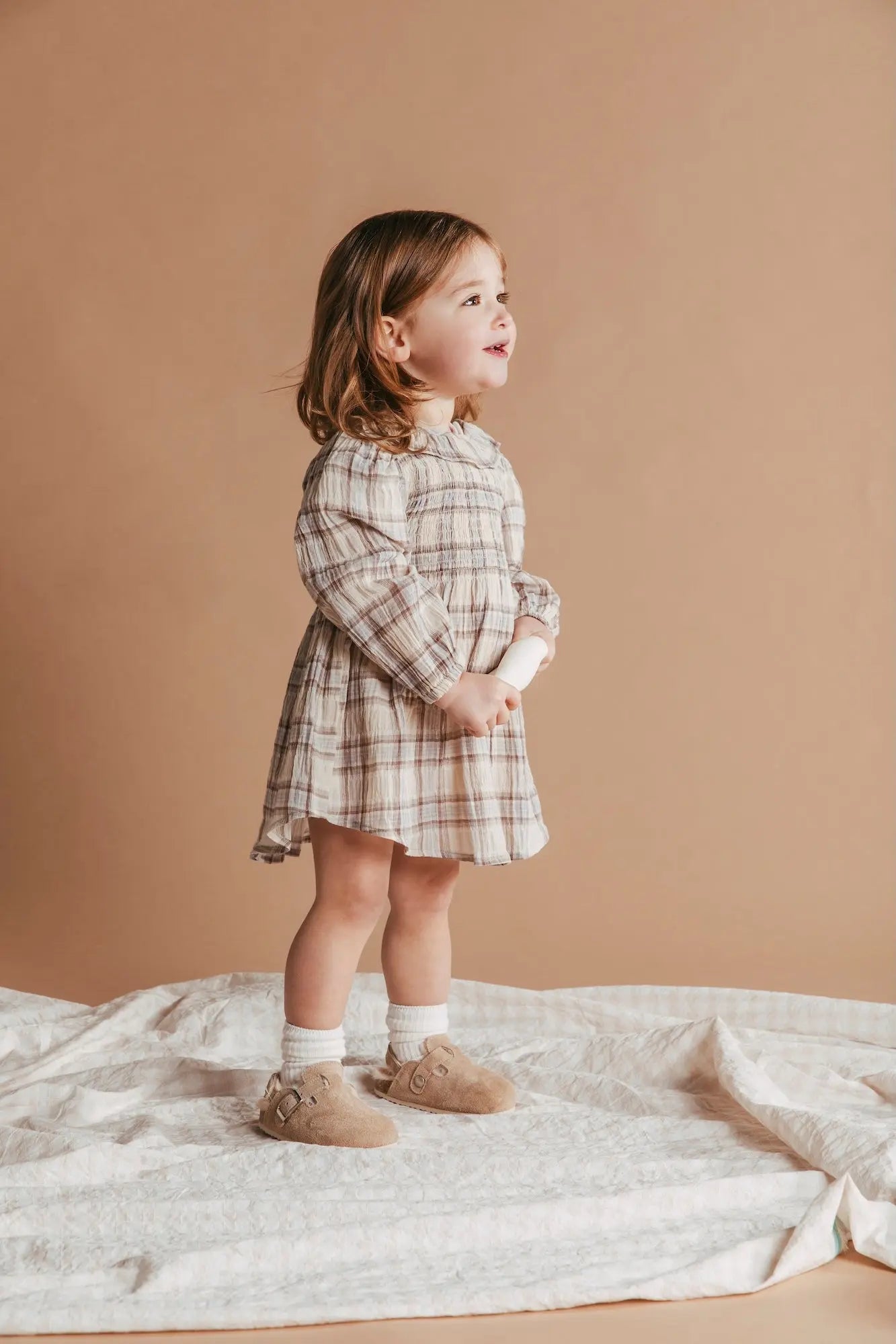 Young child wearing a plaid dress standing on a beige blanket against a beige background