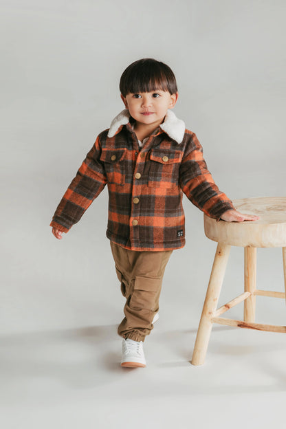 Child wearing a plaid jacket with white fur trim, standing next to a wooden stool on a plain background. Animal Crackers Standford Jacket Red Check - The Rose Edit