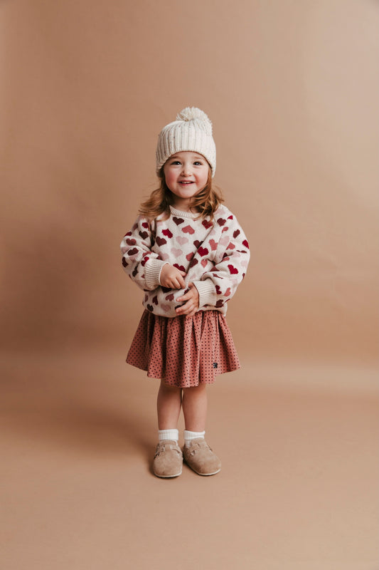 Child wearing a heart-patterned sweater and plaid skirt on a beige background