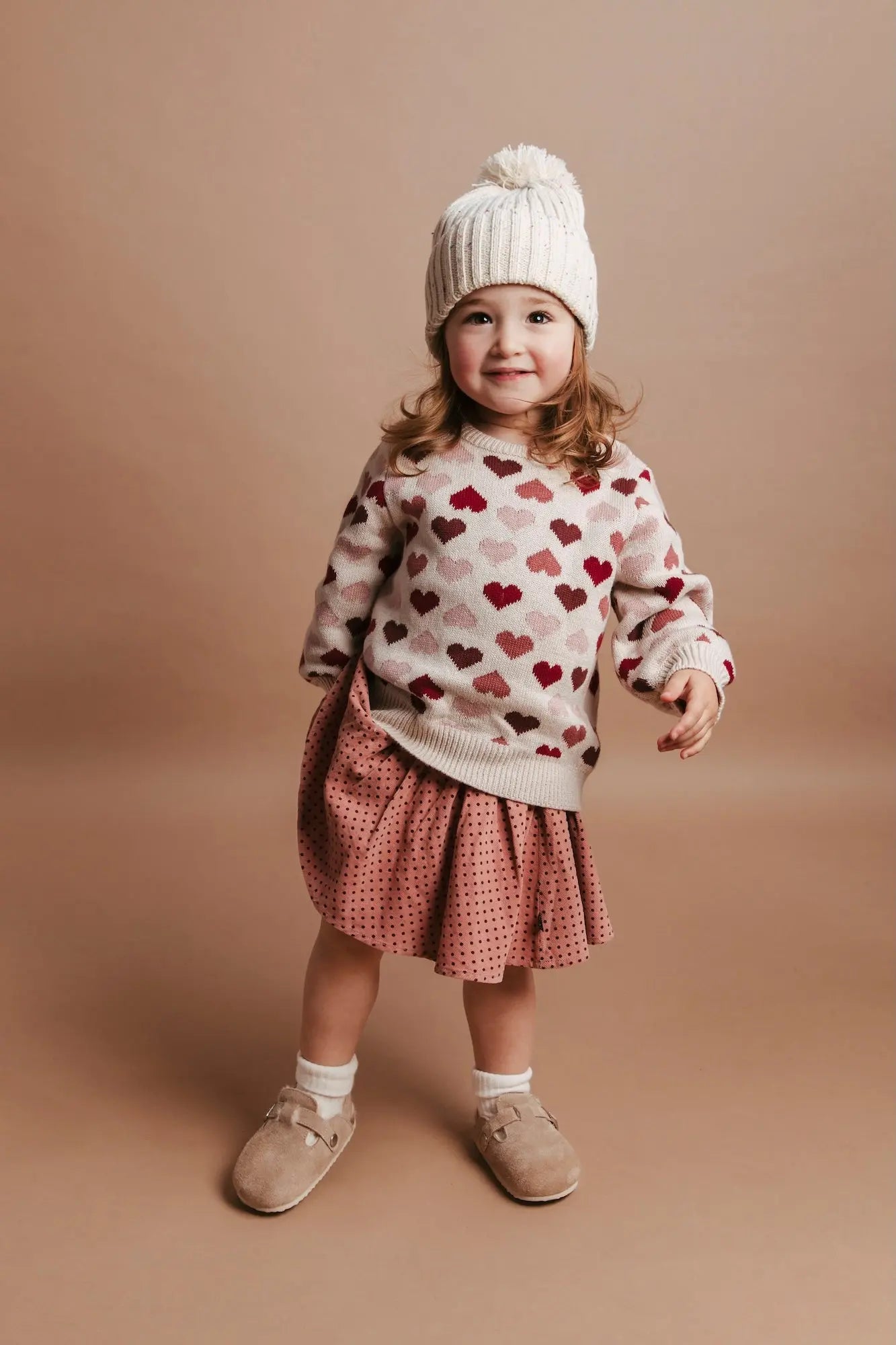 Child wearing a heart-patterned sweater and polka dot skirt on a beige background