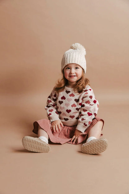 Child wearing a heart-patterned sweater and white knit hat on a beige background