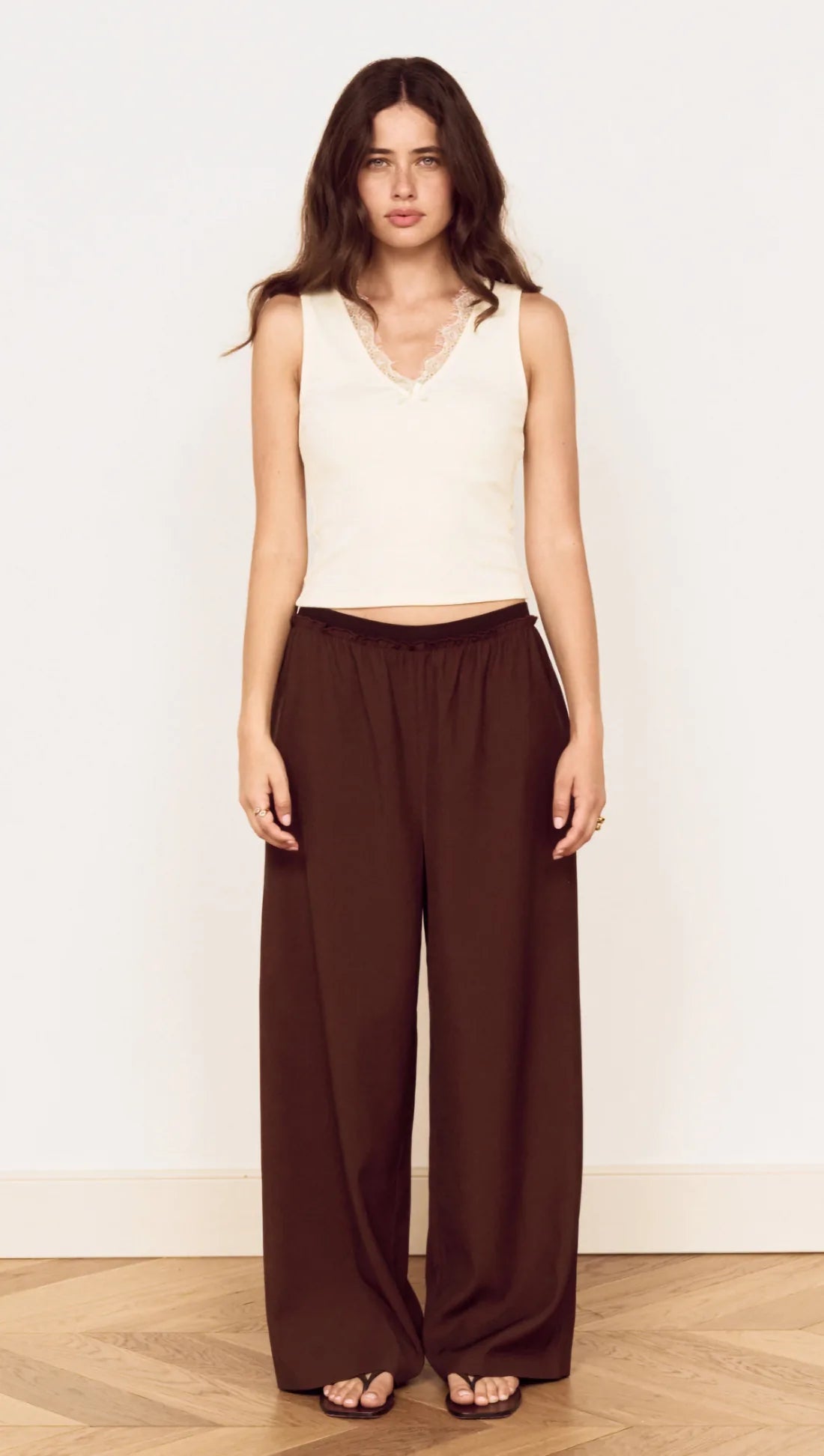 A woman modeling a sleeveless cream crop top and dark brown wide-leg trousers against a plain white wall and light wood floor.