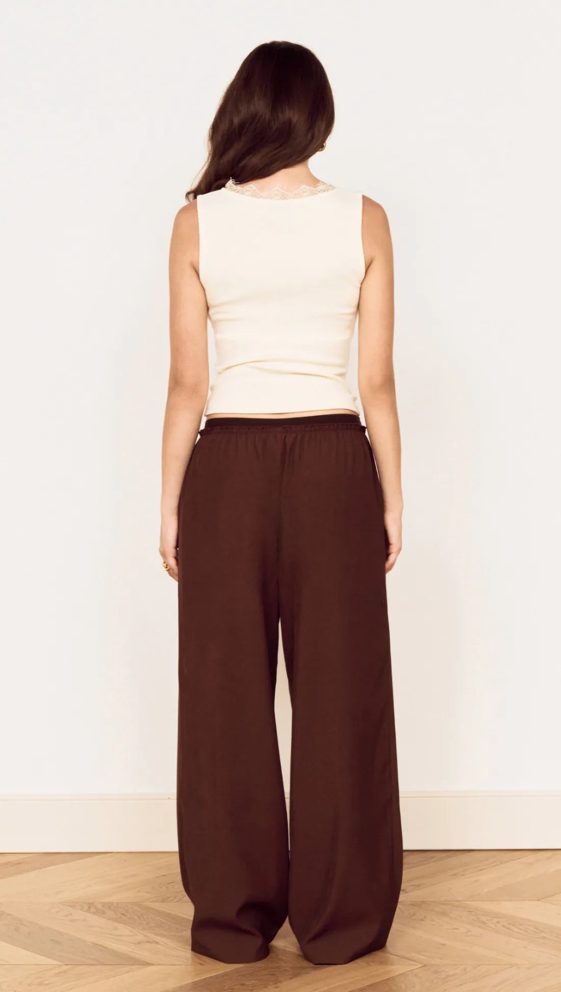 A woman shown from the back wearing a cream sleeveless top and dark brown wide-leg pants, standing in a minimalist room with a light wood floor and white wall.