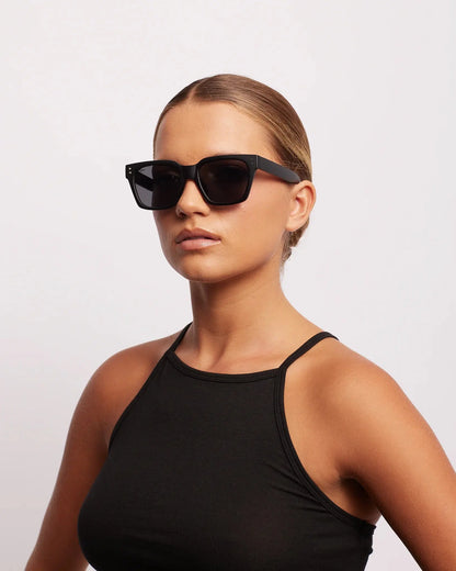 Woman wearing black sunglasses and a black tank top against a light gray background
