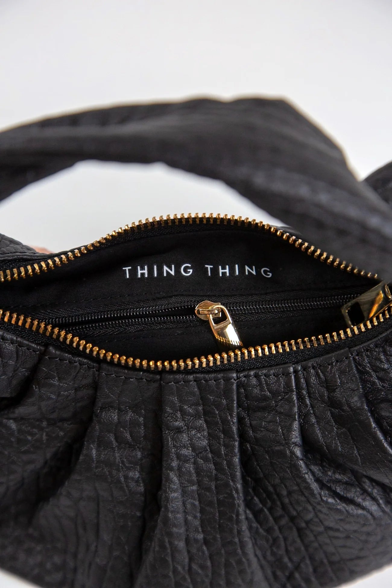 Black textured handbag with gold zipper and 'THING' branding on a white background