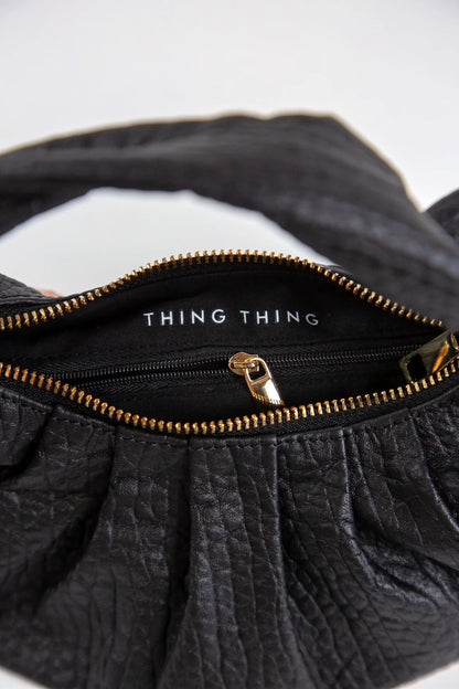 Black textured handbag with gold zipper and 'THING' branding on a white background