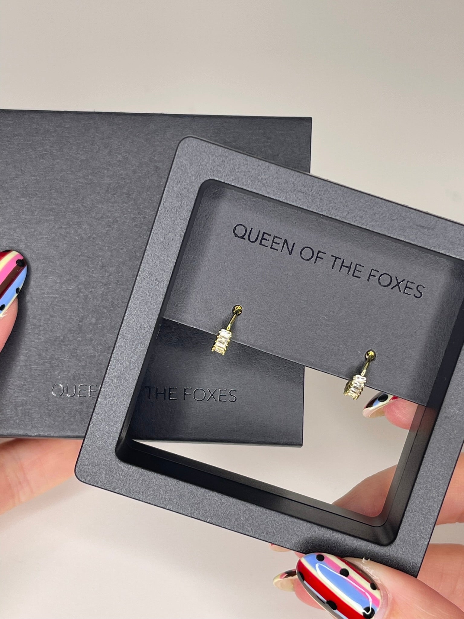 Gold earrings in a 'Queen of the Foxes' branded box held by hands with colorful nail polish. Petite Sleepers Marquise - The Rose Edit