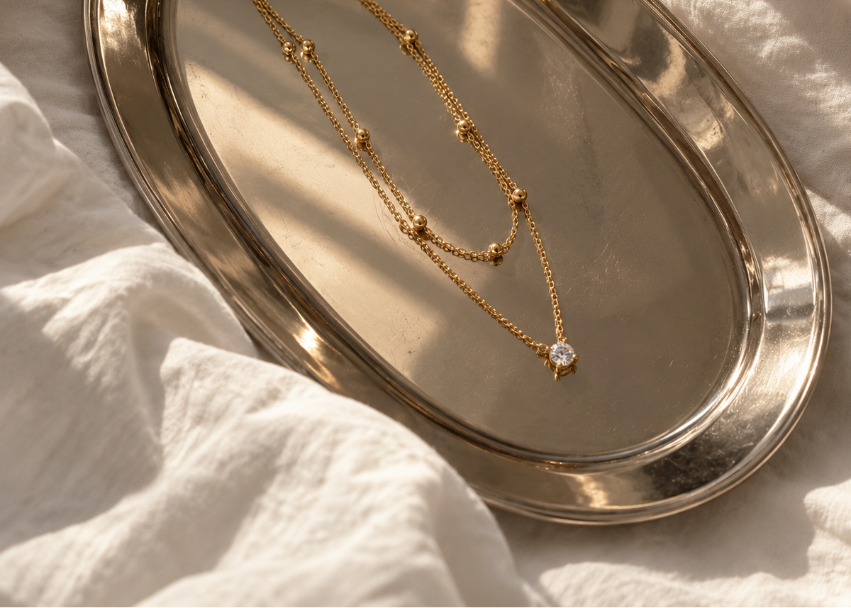 A gold-toned double-strand necklace with small bead details and a clear central stone pendant, displayed on an oval metallic tray over white fabric.