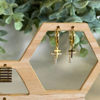Gold cross earrings on a wooden stand with green foliage in the background