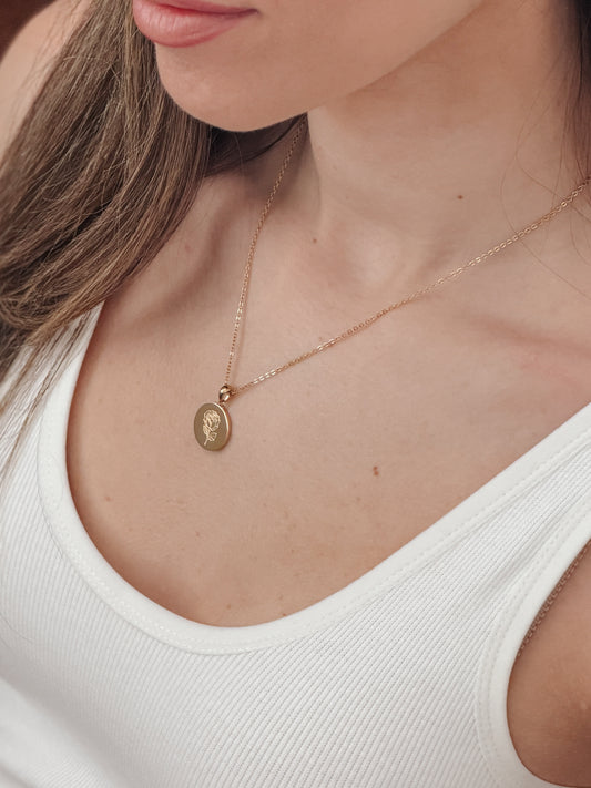 A close-up of a woman wearing a delicate gold chain necklace with an oval pendant, paired with a white ribbed tank top.