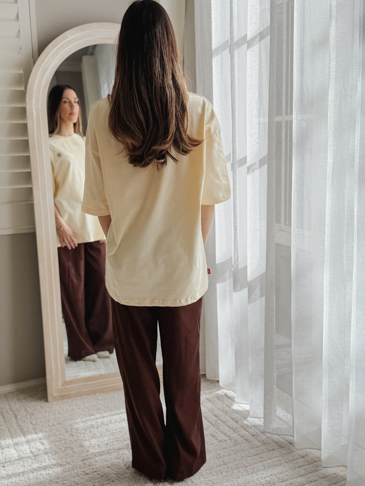 A woman stands with her back to the camera in a bright room, wearing a cream short-sleeve top and dark brown wide-leg pants. She is positioned in front of a large arched mirror that reflects her side profile, with sheer white curtains on the right.