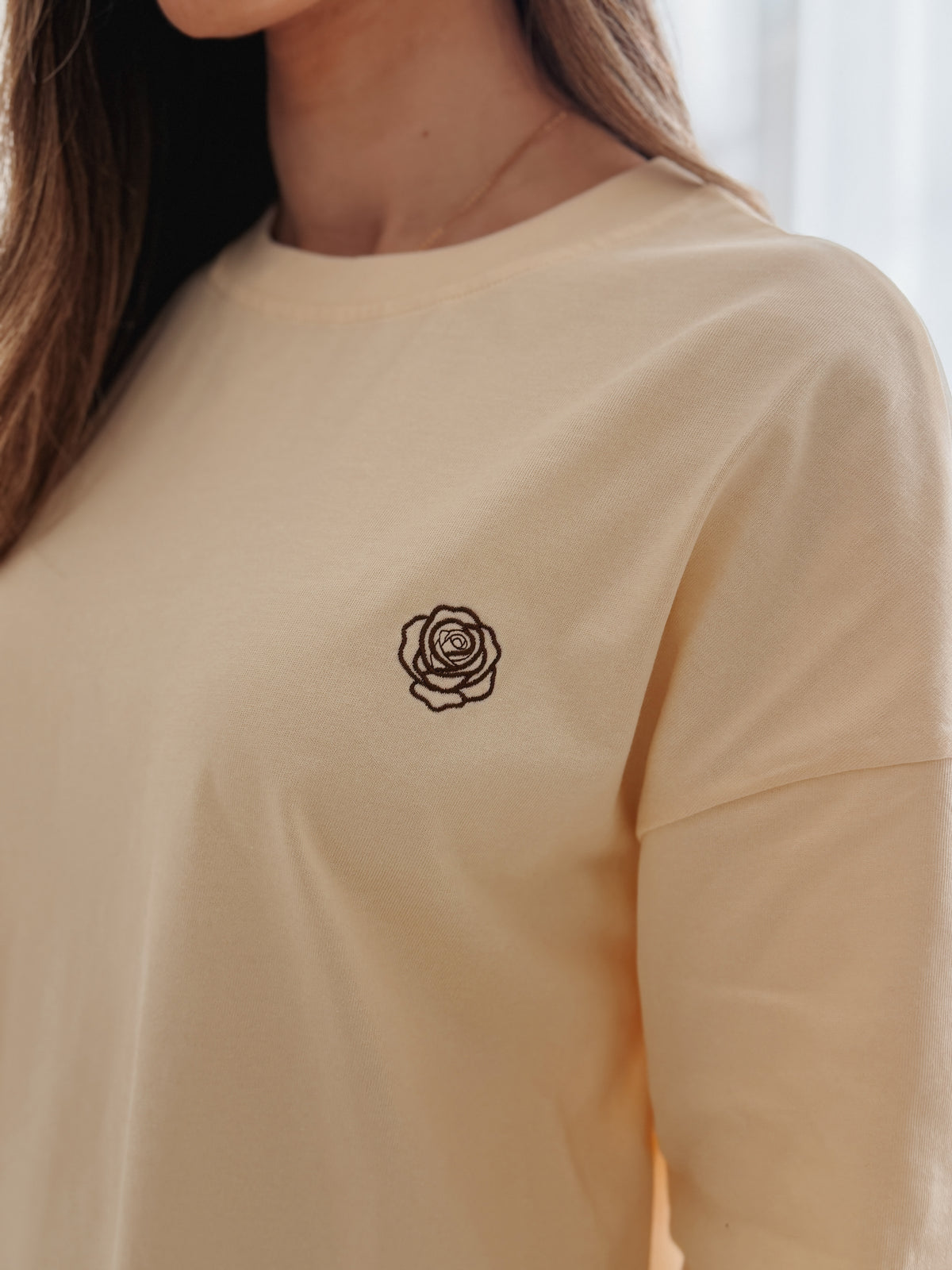 A close-up of a person wearing a light cream-colored long-sleeve t-shirt with a small black rose line-art graphic on the left chest.