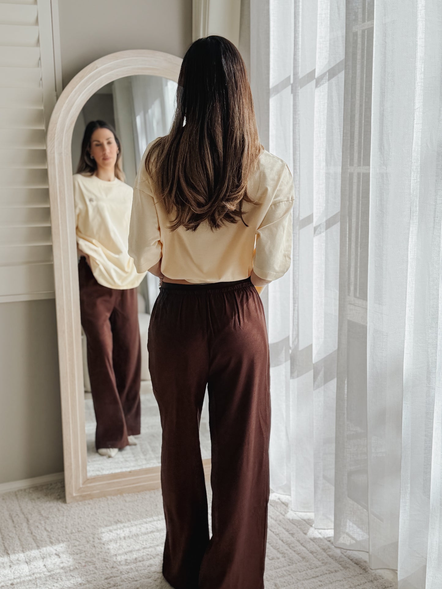 A woman stands with her back to a mirror in a bright room, wearing a cream long-sleeve blouse and dark brown wide-leg pants. Her reflection is visible in the arched mirror, with sheer white curtains and natural light on the right side.