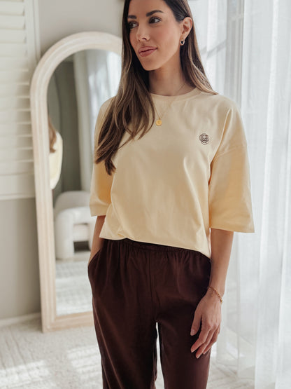 A woman wearing a pale yellow short-sleeve crewneck t-shirt with a small embroidered logo on the left chest, paired with dark brown pants, standing in a brightly lit room with a white arched mirror.