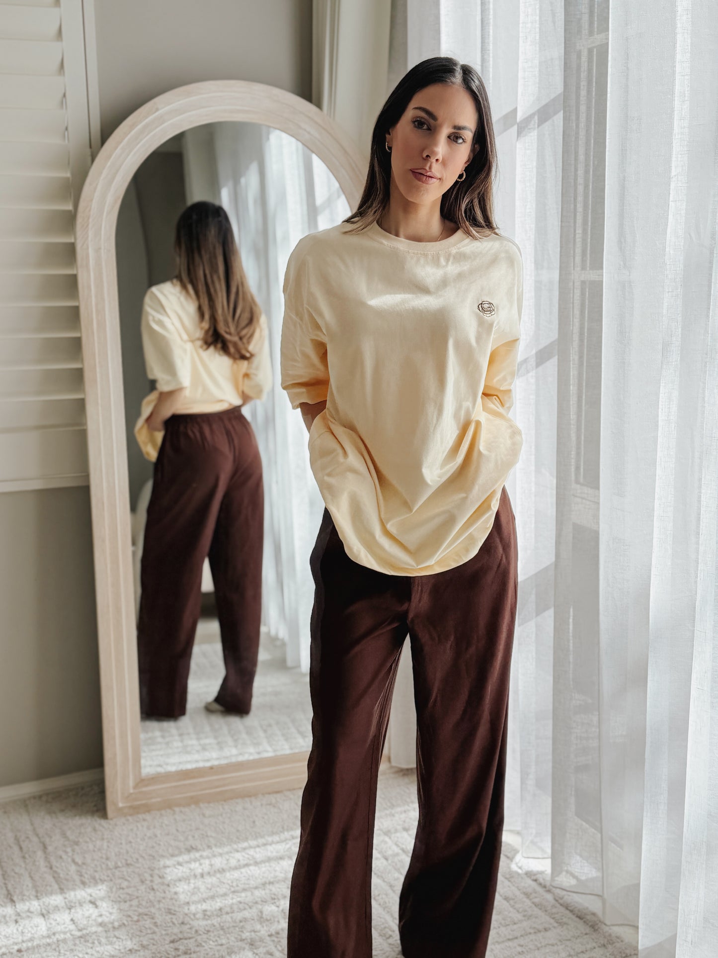A woman stands indoors near a window and mirror wearing a cream long-sleeve top and dark brown wide-leg pants. The mirror reflects her back, and sheer curtains frame the bright background.