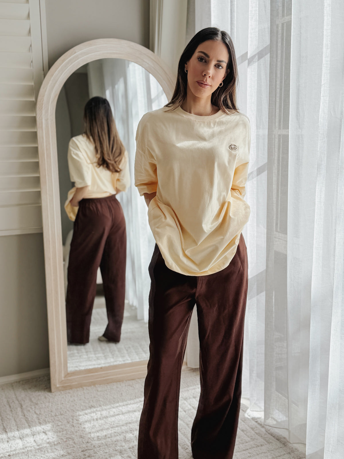 A woman stands indoors near a window and mirror wearing a cream long-sleeve top and dark brown wide-leg pants. The mirror reflects her back, and sheer curtains frame the bright background.