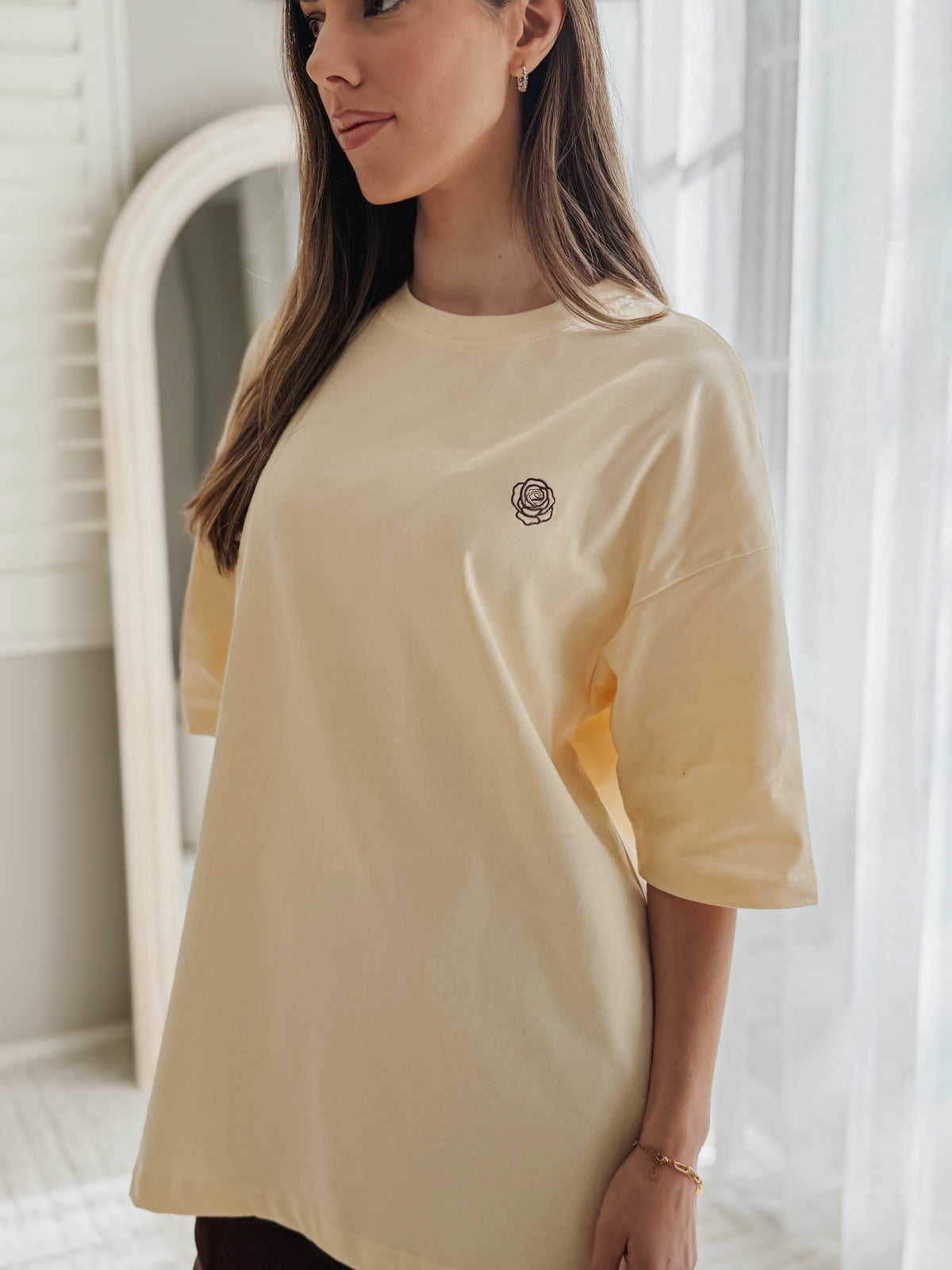 A woman wearing an oversized cream-colored t-shirt with a small embroidered rose on the left chest, standing in a brightly lit room with sheer curtains and a mirror in the background.
