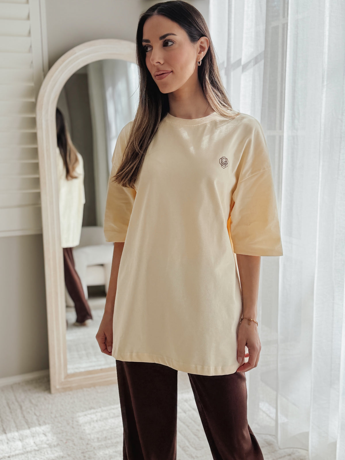 A woman wearing a cream-colored oversized t-shirt with three-quarter sleeves and a small embroidered logo on the left chest, paired with dark brown pants, standing in a brightly lit room with a mirror and sheer curtains.