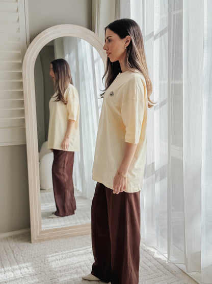 A woman stands in a bright room wearing a cream-colored short-sleeve t-shirt and brown wide-leg pants, with a large arched floor mirror behind her reflecting her side profile.