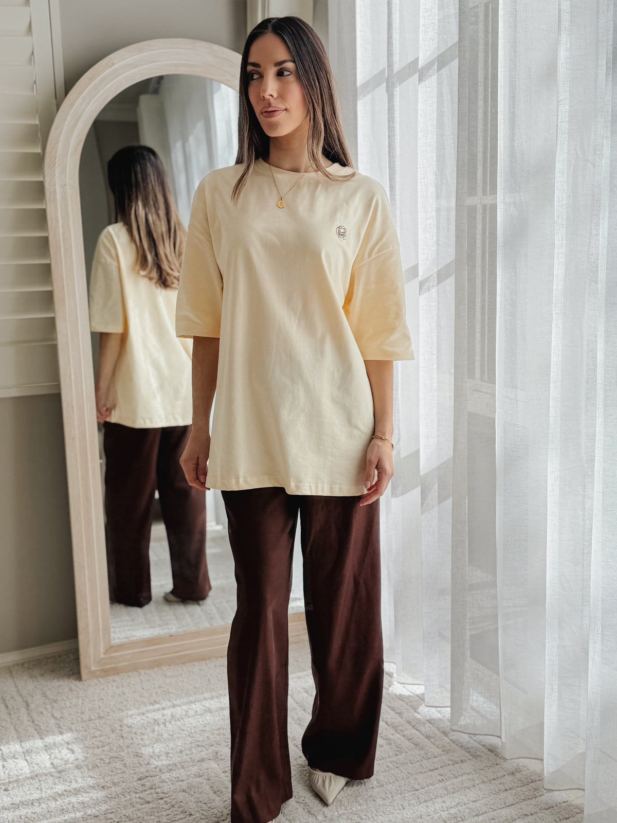 A woman stands in a bright room wearing an oversized pale yellow t-shirt and dark brown wide-leg pants, with a large arched mirror showing her reflection.