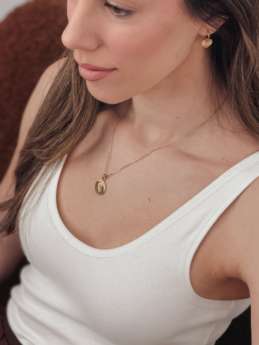 A close-up lifestyle shot of a woman wearing a delicate gold necklace with an oval pendant and a matching small gold heart earring, paired with a white ribbed tank top.
