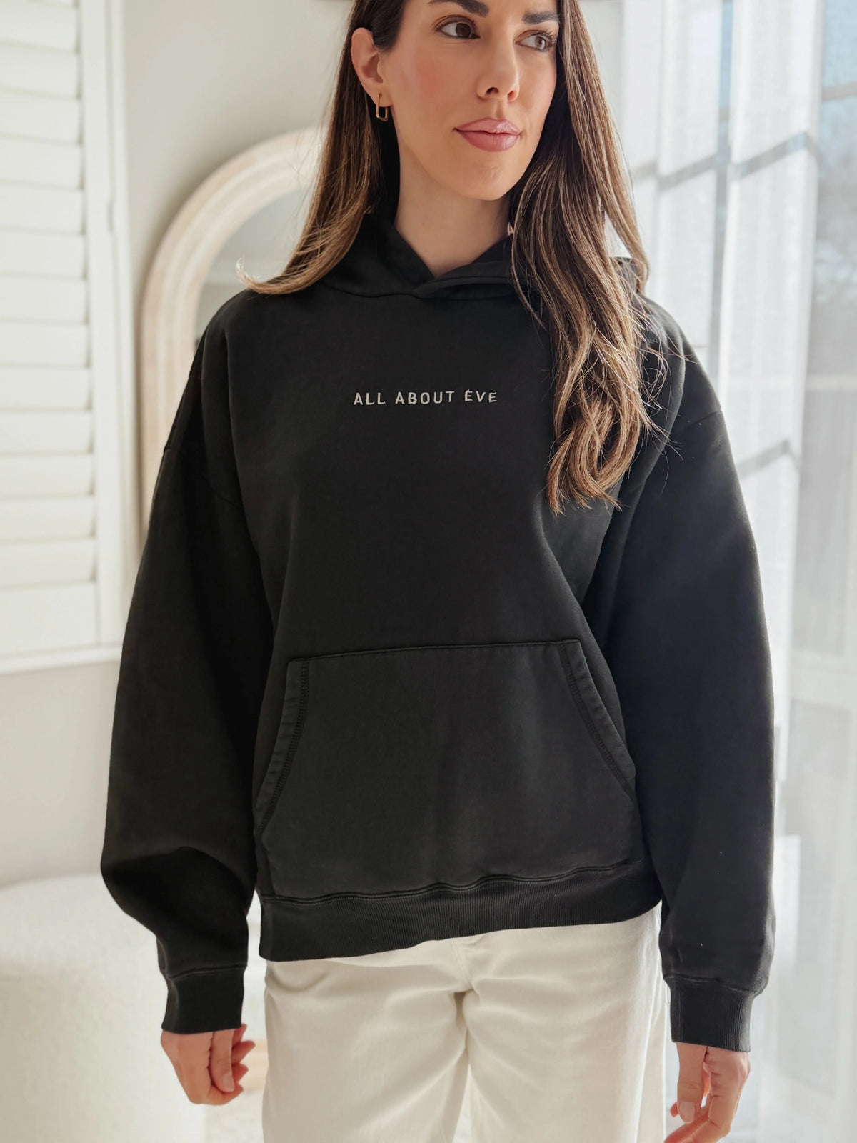 A woman wearing a black pullover hoodie with the text 'ALL ABOUT EVE' printed in white on the chest, paired with white pants, standing in a bright indoor setting.