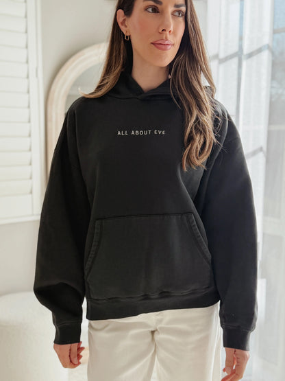 A woman wearing a black pullover hoodie with the text 'ALL ABOUT EVE' printed in white on the chest, paired with white pants, standing in a bright indoor setting.