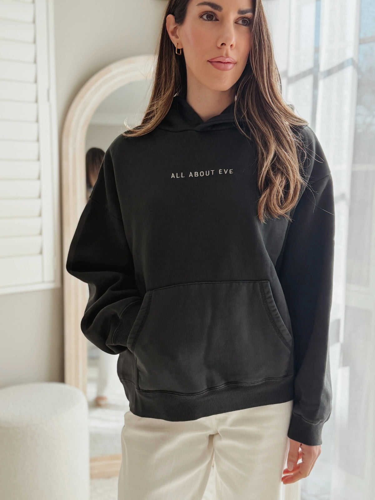 A woman wearing a black pullover hoodie with the text 'ALL ABOUT EVE' printed in white on the chest, paired with cream-colored pants, standing in a bright, modern room.