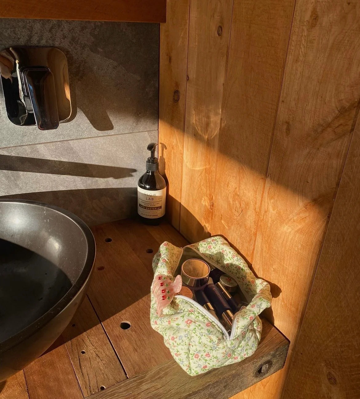 Foot with red nail polish in a floral bag on a wooden surface next to a bottle of soap. Buttermilk Grace Carry All Bag Secret Garden - The Rose Edit